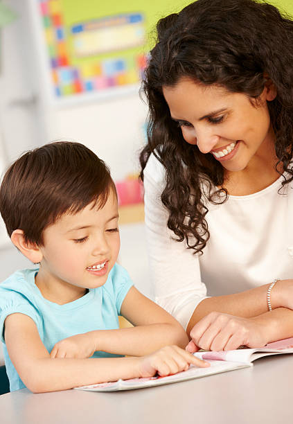 istock Elementary Age Schoolgirl Reading Book In Class With Teacher 155127534