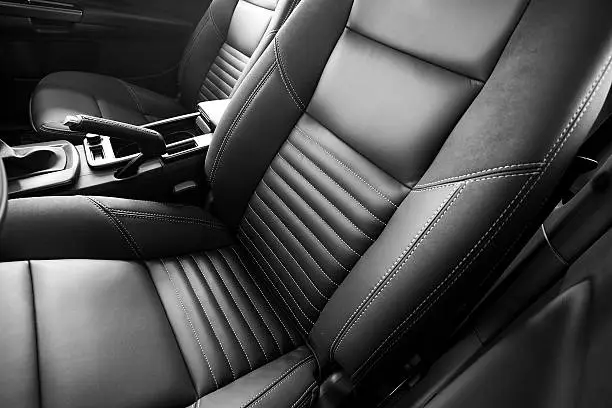 A black leather driver's and passenger's seat of a vehicle.  The two seats are divided by a center console with the emergency brake visible.  The driver's seat is positioned a bit further forward than the passenger's seat.  The seats are stitched with white thread.