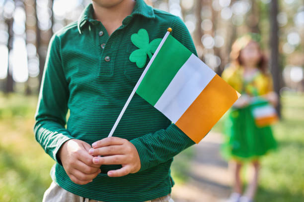 niño sosteniendo la bandera irlandesa en las manos en un feriado del día de san patricio. niño irreconocible vestido con ropa verde de duende decorada con trébol - st patricks day irish culture child leprechaun fotografías e imágenes de stock
