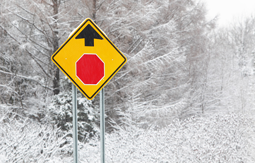 Bright yellow, red and black STOP AHEAD road sign on a rural highway during a heavy snow blizzard features internationally recognized orthographic symbols - rather than specific language words/text - to warn vehicle drivers of an imminent stop up ahead. Photo taken in Adirondacks region of northern New York State, USA.