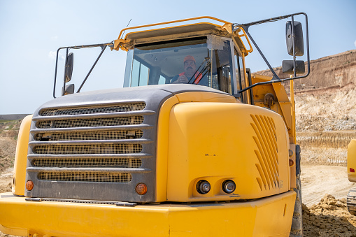 A driver of a large truck in his truck on kaolin quarry. He is wearing gray work overalls, an orange reflective vest, and an orange work cap.