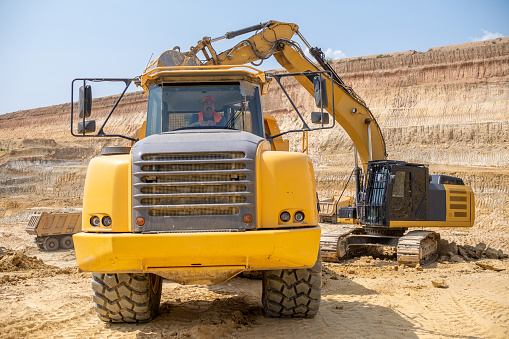 A driver of a large truck in his truck on kaolin quarry. He is wearing gray work overalls, an orange reflective vest, and an orange work cap.