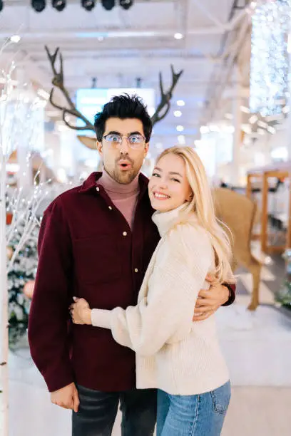 Photo of Vertical portrait of cheating wife with husband with deer antlers hanging over standing in hall of celebrate shopping mall in Christmas eve