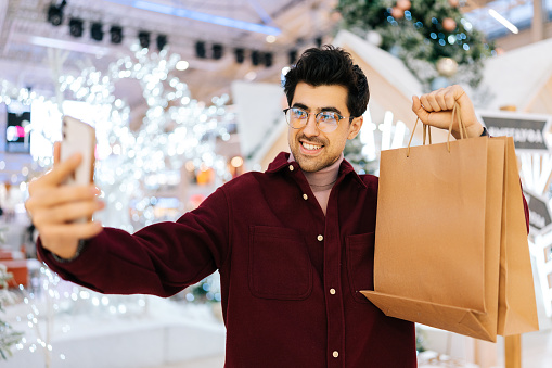 Cheerful young man holding paper bags and smiling looking at camera taking selfie on smartphone standing in hall of celebrate shopping mall in Christmas eve, on background of bright xmas decorations.