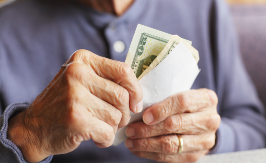 A senior man wearing a gold wedding ring is holding U.S. money and the breakfast bill/receipt while waiting to pay for his meal at a restaurant.