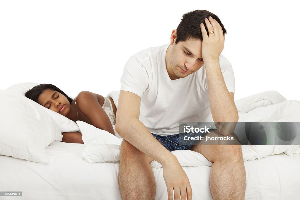 Distraught Husband Photo of an attractive young mixed-race couple in bed. Husband is sitting on the edge of the bed bed with a look of distress on his face; isolated on white. Bed - Furniture Stock Photo