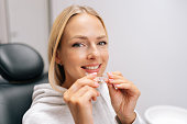Closeup portrait of smiling blonde female patient holding invisible braces aligner sitting on chair in dentistry clinic. Close up of orthodontic appliance