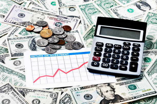 Money currency exchange coins penny dime over the hundred dollar bank notes on the white particle board table top diagonal view with vintage retro small calculator foreground defocus