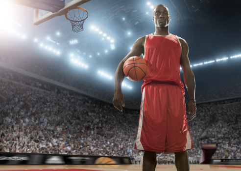 A low angle view of a professional male basketball player of African descent looking heroic, holding the basketball under his arm looking towards the camera. The athlete is standing in an empty generic indoor basketball court in an indoor floodlit arena full of spectators close to the basketball hoop. The player is wearing generic red basketball strip. 
