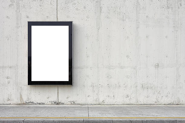 Blank billboard on wall. Blank billboard on wall. Wall is made of concrete and gray coloured. Billboard is oriented vertically and standing on the left side of frame. Edges of billboard are black. Billboard is empty so you can write or add something on it. - Clipping path of billboard included. billboard posting stock pictures, royalty-free photos & images
