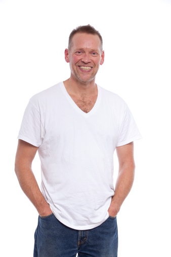 A middle aged man wearing a white undershirt and blue jeans with a bristly beard of stubble is happy and smiling about something and has a great big smile on his face.