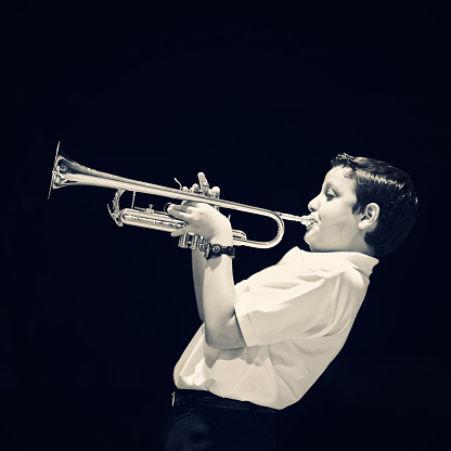 eleven years old kid playing the trumpet on black background, squared composition, desaturated.