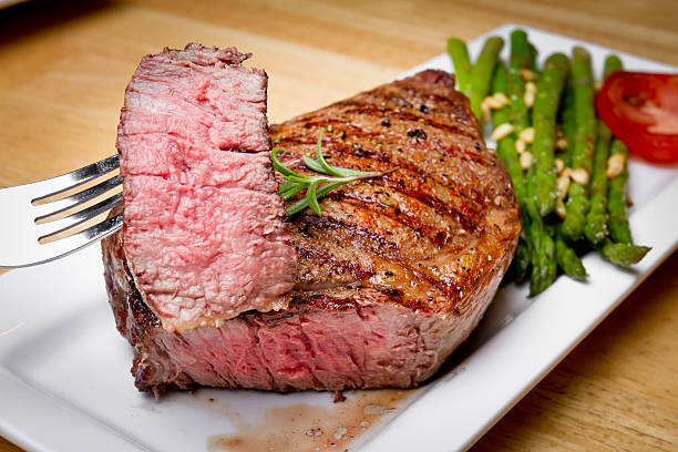 Big Rib Eye Steak With Bite Cut Out Big Rib Eye Steak  cooked medium rare With Bite Cut Out and speared on a fork with asparagus and tomato on the side.  The meal is presented on a square white plate on a light wooden table.  Steak also has a sprig of rosemary and perfect grill marks. rare stock pictures, royalty-free photos & images