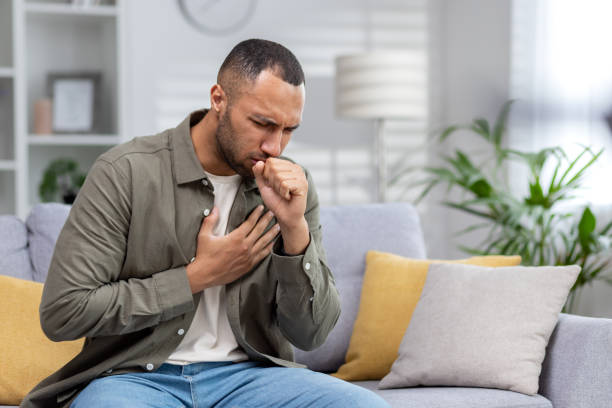 istock Young African American man sitting on the couch at home and coughing. Suffers from an attack of asthma, allergies. He holds his chest, covers his mouth with his hand 1551445607