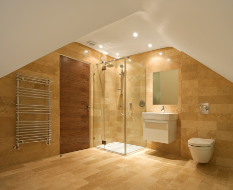 a bathroom in the attic of an expensive new home. A shower unit sits in the centre with fixed and hand shower. To the left of the solid walnut door is a modern towel radiator. To the right of the shower is a handwash basin and toilet. The walls and flooring are lined with natural stone.
