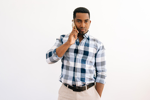 Studio portrait of confident black business man in plaid shirt talking smartphone with serious expression, having conversation on mobile phone looking at camera, standing on white isolated background.