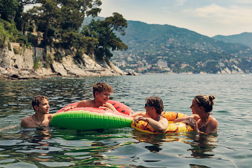 Mother and three teenage kids playing together on in the sea in Liguria, Italy.\nKids are enjoying floating in inflatables and snorkeling. In the background there are beautiful mountains and the coast of Liguria.\nShot with Canon R5