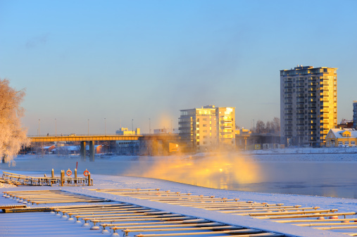 Bridge and buildings along riverbank on a cold winter day. Mist rising from freezing water. Scene lit by low angle sun.