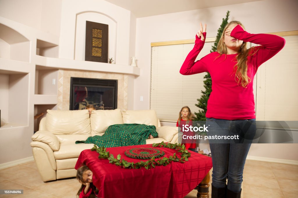 Babysitter does Holiday Countdown to Chase of Hide Holiday themed view of college aged babysitter stands with her eyes covered and two fingers showing only 2 seconds till the game begins, waiting for start has children bored to hide in their Christmas setting of a Southwestern style living room 10-11 Years Stock Photo