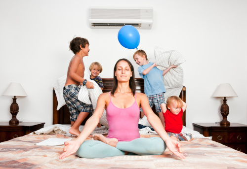 A mother in the lotus position meditating while children jump and play around her.