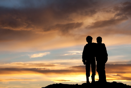 Two happy young boys on a hill at sunset. Back view. Children are elementary aged and have their arms around each other in embrace. Unrecognizable models. Themes in the image include best friends, love, relationships, siblings, brothers, bonding, care, comfort, talking, outside, nature, recreation, leisure, watching the sunset, beauty, and family. 