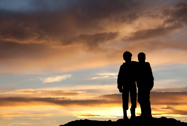 Happy Children Watching Sunset Two happy young boys on a hill at sunset. Back view. Children are elementary aged and have their arms around each other in embrace. Unrecognizable models. Themes in the image include best friends, love, relationships, siblings, brothers, bonding, care, comfort, talking, outside, nature, recreation, leisure, watching the sunset, beauty, and family.  arm in arm stock pictures, royalty-free photos & images