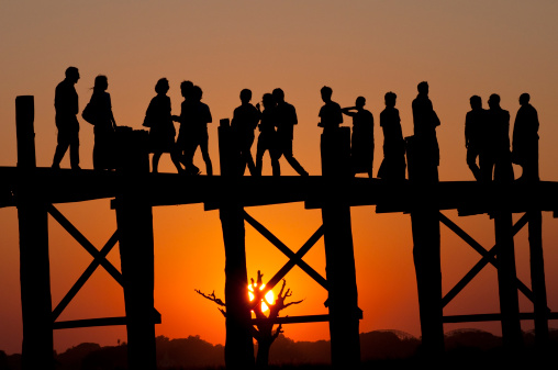 Fifteen people of various ethnicities and both genders standing on a bridge at sunset. Location: U Bein Bridge in Amarapura, Myanmar