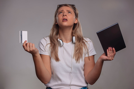 Shocked woman holding credit card and tablet computer isolated on a gray background