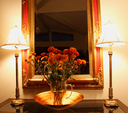 Bouquet of Bronze Chrysanthemums in front of gilded mirror at night. Two delicate slim bronze lamps on either side. All sitting on glass top table.
