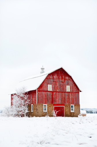 A old red barn with weathered siding and stone walls. There is fresh snow on the roof and grounds.