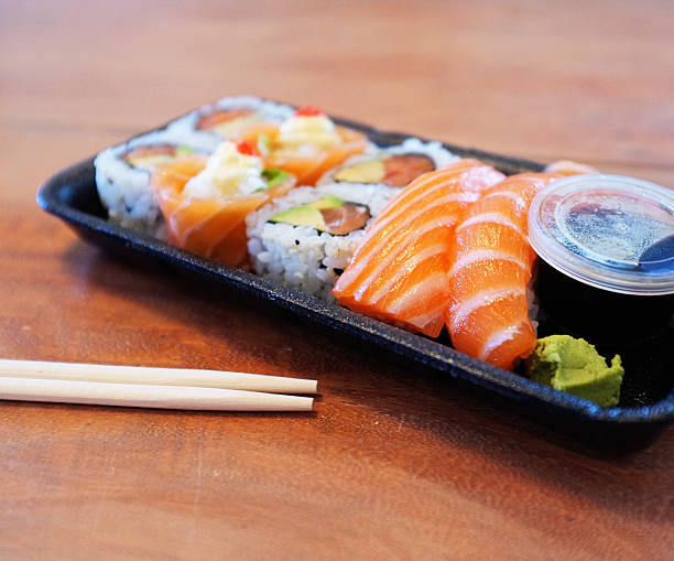 A sushi take-out containing a selection of salmon sushi specialities and sauces in a polystyrene tray with chopsticks on the side sits ready and waiitng on a wooden table. 