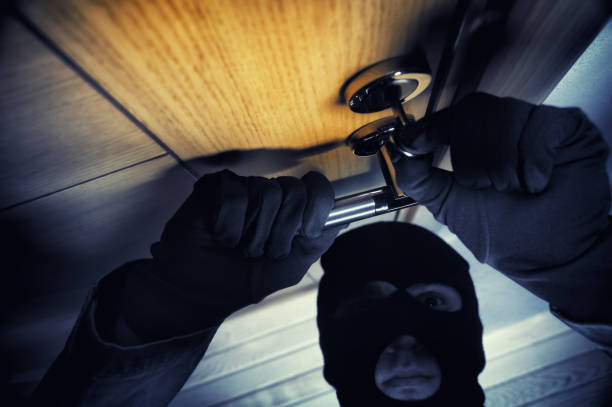 A person in a dark hoodie and black ski mask looks intensely at the camera while partially concealing their face and holding a crowbar. The shattered glass background and dramatic lighting emphasize themes of intrusion, secrecy, and criminal activity. This photo is suitable for crime, law enforcement, and urban security stories.