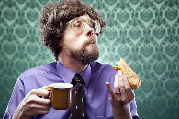 A vintage dressed man with large glasses and bad hair in a 1970's styled office enjoys a coffee break with a maple bar donut.  Horizontal with copy space on damask print wallpaper.