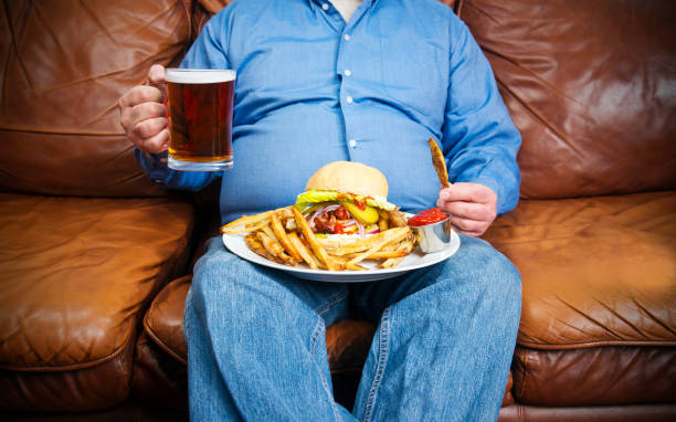 Overweight man over-eating on a couch An overweight man over-eating on a couch. High glycemic index foods. pot belly stock pictures, royalty-free photos & images