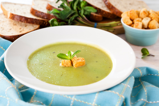 Pea and mint soup topped with croutons and fresh mint, shown on a white table with ingredients.
