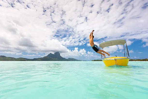 Young boy jumping from yellow motor boat into the waters of a turquoise lagoon. Beautiful cloudscape and clear turquoise lagoon. Bora Bora Lagoon, Society Islands, French Polynesia.