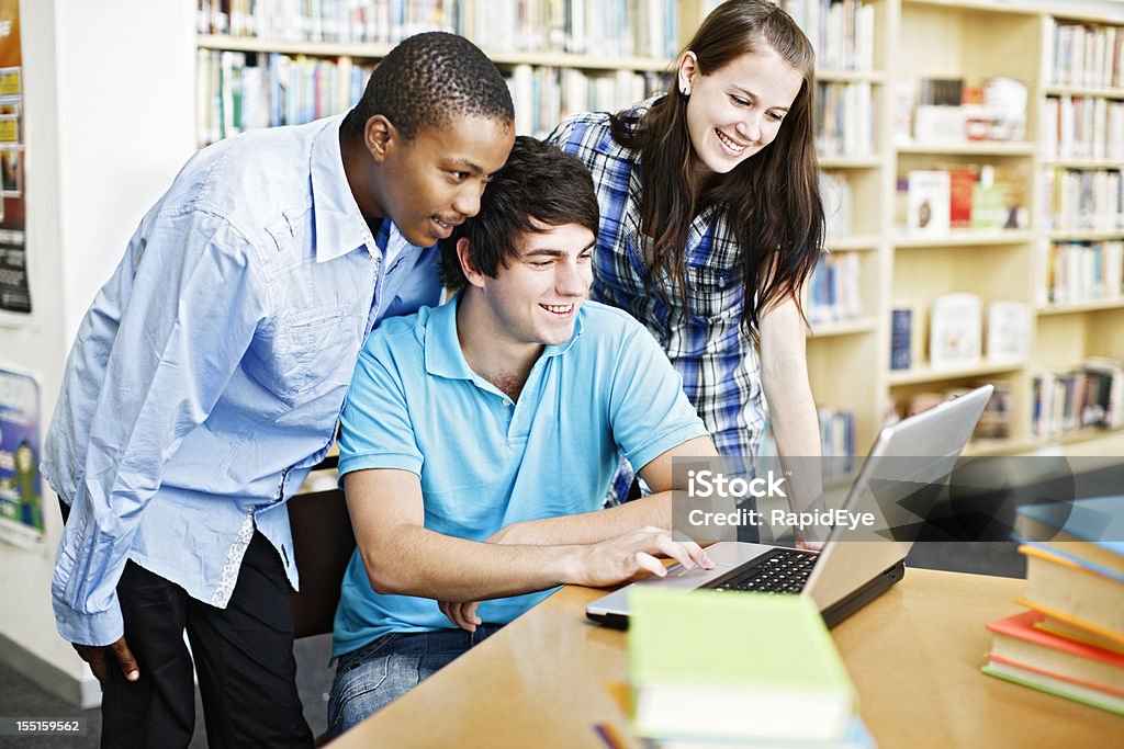 Trois étudiants heureux sur un ordinateur portable dans la bibliothèque - Photo de Adulte libre de droits