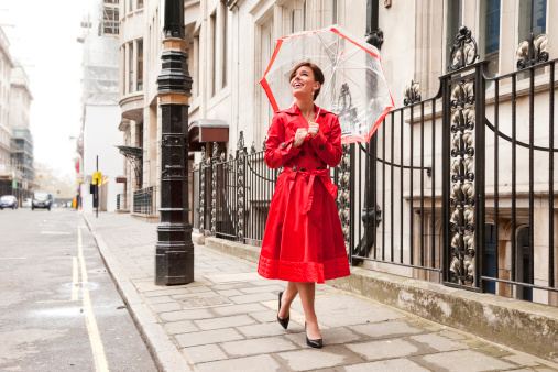 Photo of a young woman in a bright red trench coat, walking along a London sidwalk with an umbrella and a bright smile; slight motion blur from walking activity.
