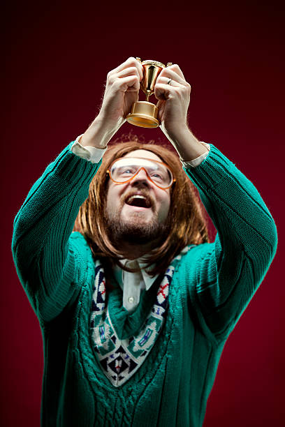 Goofy Nerd With Bad Sweater Wins Trophy A long haired nerd man with beard, long hair, funny glasses, and ugly sweater holds up a medal or trophy that he won with joy and excitement on his happy face.  Vertical. nerd sweater stock pictures, royalty-free photos & images