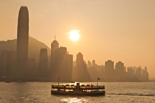 Warm orange light from the setting sun silhouetting the iconic skyscrapers and crowded cityscape of Hong Kong Island above the famous Star Ferry plying its trade from Kowloon across Victoria Harbour, China. ProPhoto RGB profile for maximum color fidelity and gamut.