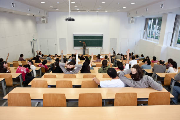 Classroom with multiple students sitting at long tables in wood-backed chairs.  The walls in the classroom are white, and the ceiling has multiple cylinder lights.  There are two sets of windows on the far right side of the classroom.  A male professor stands in front of the classroom and writes on a dark green chalkboard with a piece of chalk, and the students appear to be in their teens and 20s.  A white line is drawn through the top portion of the chalkboard.  Two white loudspeakers hang from the ceiling on the right and left sides of the room.