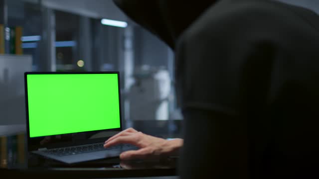 Close up of hacker in hoodie working on laptop with green screen in dark office