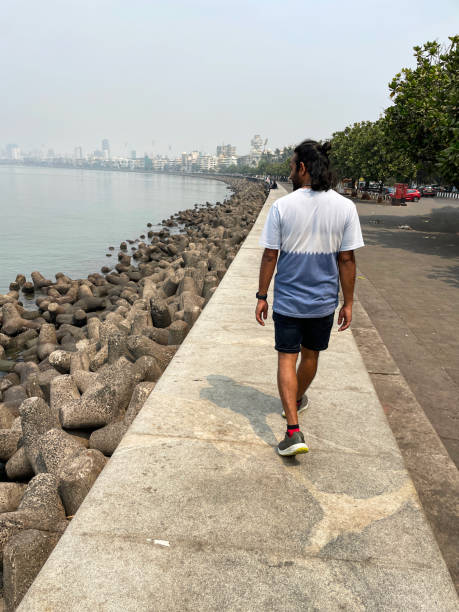 Stock photo showing close-up view of a manmade, sea defence of a concrete, seawall, which has been constructed to prevent flooding of the promenade of Marine Drive, Mumbai, India during high tide. A stack of large, concrete, tetrapod blocks, which have been piled up to provide a retaining wall to protect the promenade is protecting the roadway by helping to reduce the sprays and splashes from the sea waves. imagen en primer plano de un hombre caminando sobre el malecón marine drive, mumbai, india, defensa de hormigón, gestión costera, prevención de la erosión, agua de mar, paseo en forma de c, edificios art deco y rascacielos que bordean la playa públic
