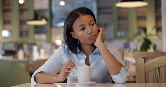 Bored businesswoman drinking coffee waiting for colleague in restaurant. Portrait of african sad female sitting alone in cafe waiting for date