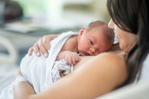 A new Mother holds her baby gently to her chest as they spend time bonding with some skin-to-skin time.  She is wearing hospital gown and the baby is swaddled loosely as Mom and baby spend time together shortly after delivery.