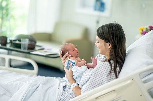 A new Mother sits up in her hospital bed shortly after delivery as she holds her newborn out in front of her and studies his features.  She is wearing a hospital gown and is laying in her hospital bed with the inafnt.
