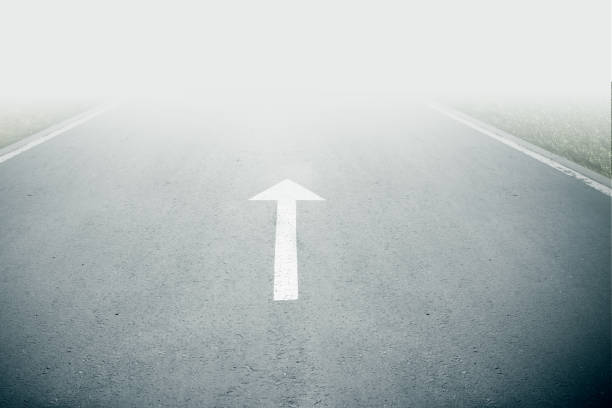White arrow sign on road goes into fog. Direction way sign. Moving forward concept. Unclear ahead or negative tomorrow concept. Empty copy space.