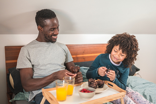 Waist up shot of a mid adult Black man  and his  cute son with curly hair in pajamas, sitting on the modern bed  and eating muffins and fruit from a foldable table, having fun.