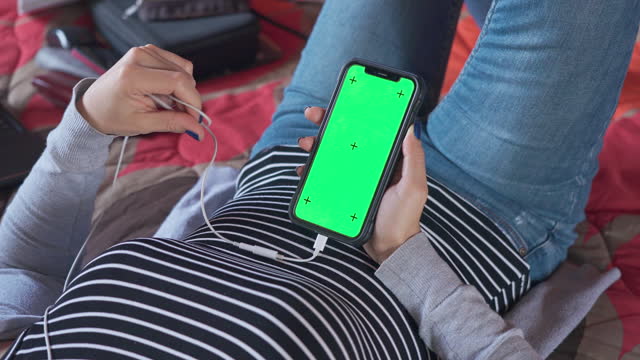 Hispanic young woman listening to music on smartphone with chroma key blank screen while laying on a bed