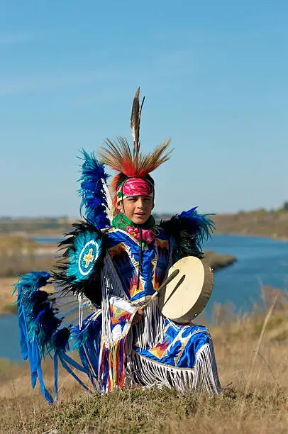 Boys Fancy Dancer with hand drum posing in a prairie field with a river background